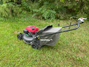 A Honda walk-behind lawn mower with a red engine cover and a black grass bag, positioned on a grassy lawn with bushes in the background.