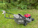 A Honda walk-behind lawn mower with a black handle and grass bag, featuring a red engine cover, resting on a grassy lawn with green bushes in the background.