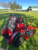 A red and black stand on lawn mower with a rear operator station, photographed outdoors on a green grass lawn with trees and a building in the background.