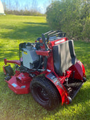 A red stand-on lawnmower with a grey knee pad and black tires, photographed outdoors on a grassy lawn with trees in the background. WIth ICD cutting systems label on the deck.