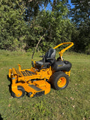 A yellow and black zero-till riding lawn mower with a high arm, parked on a grassy lawn with trees in the background.